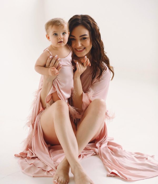 Smiling mommy with newborn baby in same pink dresses posing in the studio on white background.  Close up photo of mother in pink stylish dressing gown playing with her 9 month old baby girl. Copy space for text